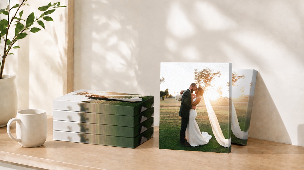 Wedding photo canvas prints displayed beside a stack of canvases on a sunlit wooden table.